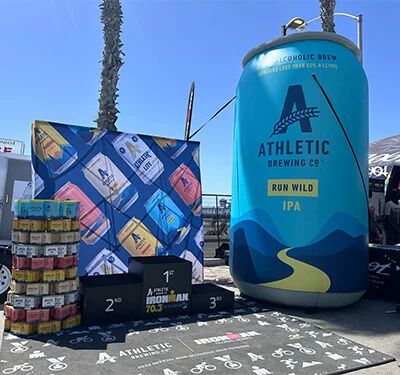 A large inflatable can of Athletic Brewing Co. Run Wild IPA stands next to a backdrop with colorful Athletic Brewing cans, a stack of real cans, and podium blocks for a race under sunny skies.