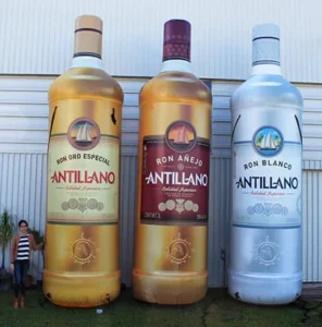 A woman stands next to three giant inflatable bottles of Antillano rum—one gold, one brown, and one silver—displayed outdoors against a white building wall.