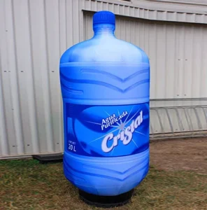 A large outdoor advertisement shaped like a blue 20-liter water bottle with the label Agua purificada Cristal, standing on grass near a corrugated metal building.