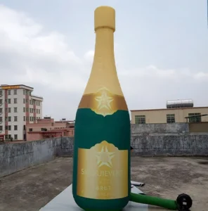 A large inflatable champagne bottle stands upright on a rooftop, with buildings and a cloudy sky in the background. The bottle is green and gold with a star logo and the word SIMAJUEVENT on it.