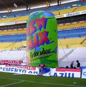 A large inflatable can advertising New Mix stands on a soccer stadium field near the stands, with several people nearby and colorful stadium seats in the background.