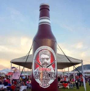 A giant inflatable bottle of Cerveza Zapata stands at an outdoor festival, with crowds of people, tents, and mountains visible in the background at sunset.