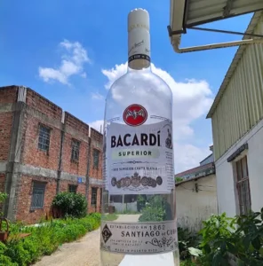 A large Bacardi Superior rum bottle appears to stand upright outdoors between two industrial buildings, but it’s an optical illusion created by angling a normal-sized bottle close to the camera. The sky is blue with clouds.