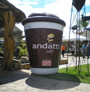 A giant inflatable coffee cup with andatti café and the OXXO logo stands outdoors on a sunny day, surrounded by people, tents, trees, and a rustic wooden structure.