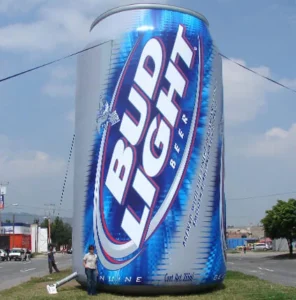 A person stands next to a huge inflatable Bud Light beer can on a grassy area near a road, making the can appear much larger than life. Buildings and trees are visible in the background.