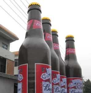 Five large novelty beer bottle statues with Budweiser labels are standing outdoors near a building and power lines, viewed from a low angle against a cloudy sky.