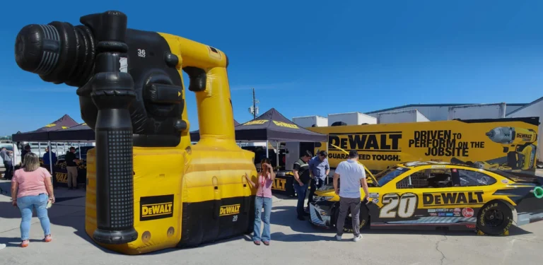 People stand near a giant inflatable DeWalt power drill and a yellow DeWalt-themed race car, with branded tents and banners in the background, at an outdoor event under a clear blue sky.