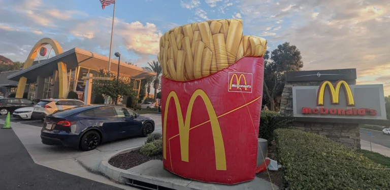 A giant inflatable McDonalds French fries display stands outside a McDonalds restaurant at sunset, with a car parked nearby and the restaurants golden arches sign visible.
