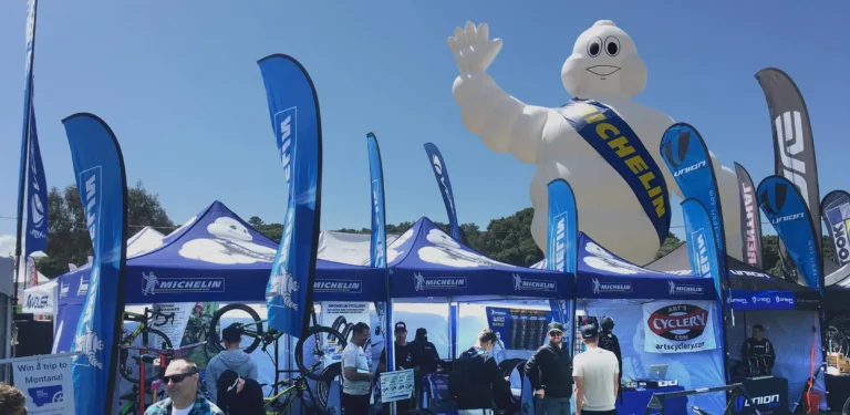 A large inflatable Michelin Man looms over a busy outdoor event booth with blue tents and flags displaying the Michelin logo. People are gathered around, some with bicycles, under a sunny blue sky.