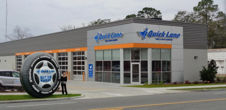 A Quick Lane Tire & Auto Center building with large windows and an orange accent stripe. A person stands beside a giant tire display advertising a sales event near the entrance.