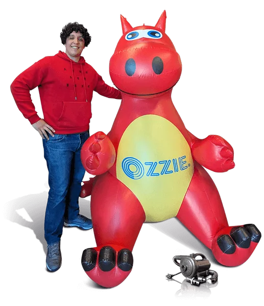 A person in a red hoodie stands smiling next to a large red inflatable cartoon dragon with a yellow belly labeled OZZIE. A small metallic object sits on the ground in front of them.