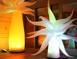 Two large, glowing, spiky inflatable sculptures—one yellow-orange, one white with hints of blue and green—stand indoors near glass doors, brightly illuminating the area.