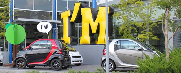 Two small Smart cars parked outside a modern glass building, with a large yellow and black IM sign and a speech bubble reading IM! behind them. Trees and shrubs are visible in the foreground.
