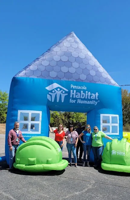 Five people stand in front of a large inflatable blue house with Pensacola Habitat for Humanity written on it, and two large green inflatable objects shaped like snails on each side. The sky is clear and sunny.