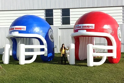 A person stands between two large inflatable football helmets, one blue and one red, on a grassy area in front of a white building. The helmets display a National Guard logo on the front.