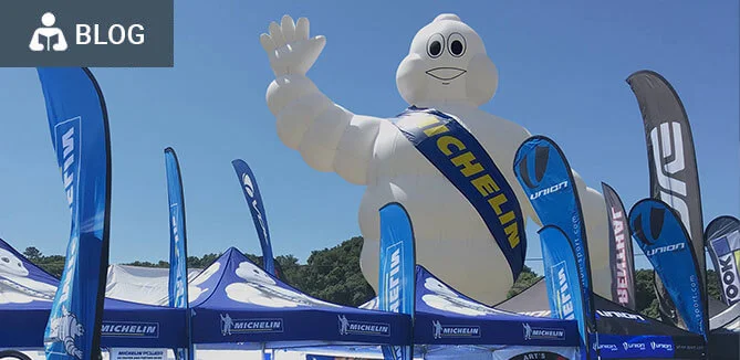 A large inflatable Michelin Man with a blue sash stands among blue Michelin flags and tents at an outdoor event under a clear blue sky.