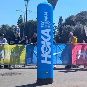 A tall, blue inflatable pillar with the HOKA logo and slogan FLY HUMAN FLY stands in front of a race barrier, with spectators and colorful banners in the background.