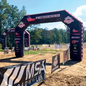 An outdoor event entrance arch reads #MEETUSINTHEMUD with various sponsor logos, set on a dirt track surrounded by trees and banners under a clear sky.