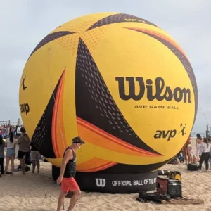 A giant inflatable Wilson AVP volleyball is displayed on a sandy beach, surrounded by people. A man in a tank top and hat walks in the foreground. The beach is busy with spectators in the background.