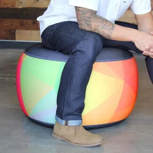 A person in jeans and brown shoes sits on a round, colorful ottoman with a geometric rainbow pattern. Only the lower half of their body and one tattooed arm are visible.