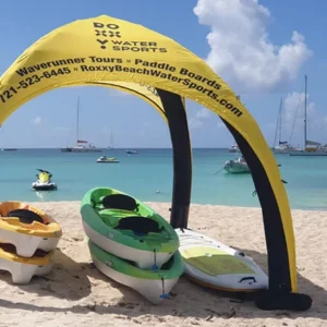 Yellow and black canopy labeled “DO IT WATER SPORTS” on a sandy beach, with kayaks and paddleboards beneath it. Clear turquoise water, sailboats, and yachts are visible in the background under a blue sky.
