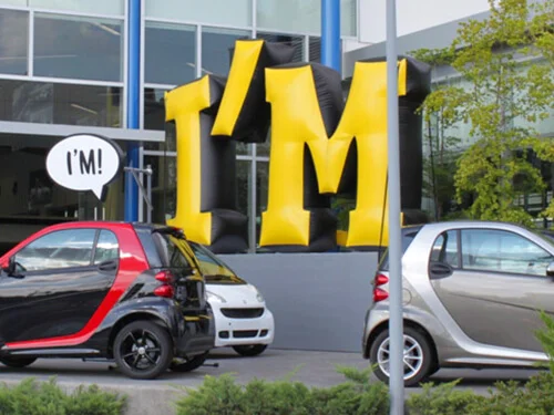 Two small cars are parked in front of a building with large, inflatable yellow and black letters spelling IM. A speech bubble next to one car also says IM! There are trees and glass windows in the background.