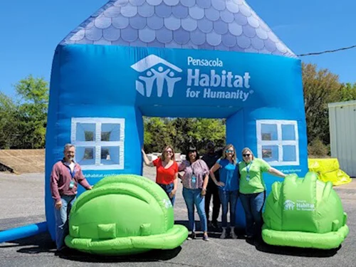 Five people stand smiling in front of a large inflatable blue house with Pensacola Habitat for Humanity on it. Two large green inflatable hard hats are on the ground beside them. Trees and a building are in the background.