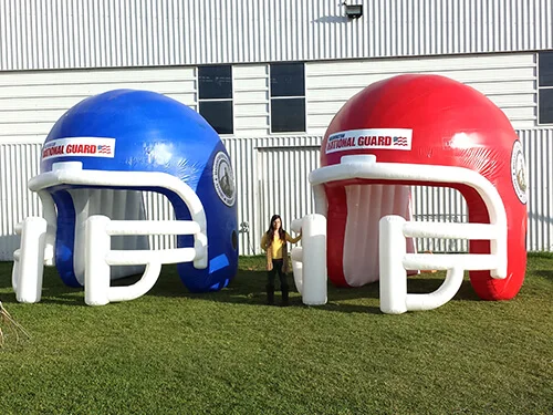 A person stands between two giant inflatable football helmets, one blue and one red, on a grassy area outside a white industrial building. The helmets have “National Guard” written on them.