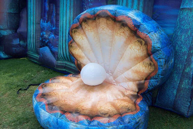An inflatable clam shell with a large white pearl inside sits on grass, surrounded by an underwater-themed background with blue columns and seaweed designs.