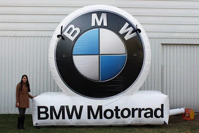 A woman stands next to a large inflatable BMW logo with the words BMW Motorrad printed on its base, placed outdoors in front of a metal building.