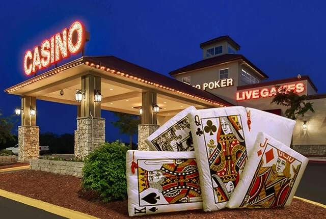 A brightly lit casino building at night with signs reading CASINO, POKER, and LIVE GAMES. In the foreground, large playing cards with royal faces are displayed over landscaping.