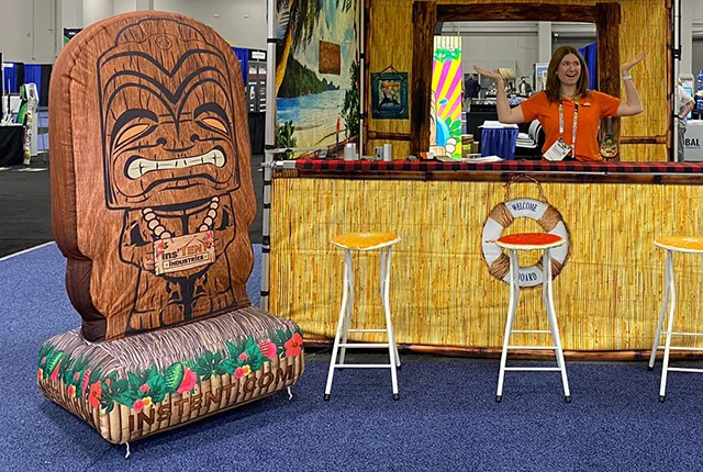 A woman in an orange shirt stands behind a tiki-themed booth with a bamboo counter, two stools, and a large inflatable tiki statue on the left. The scene is decorated to resemble a tropical island bar.