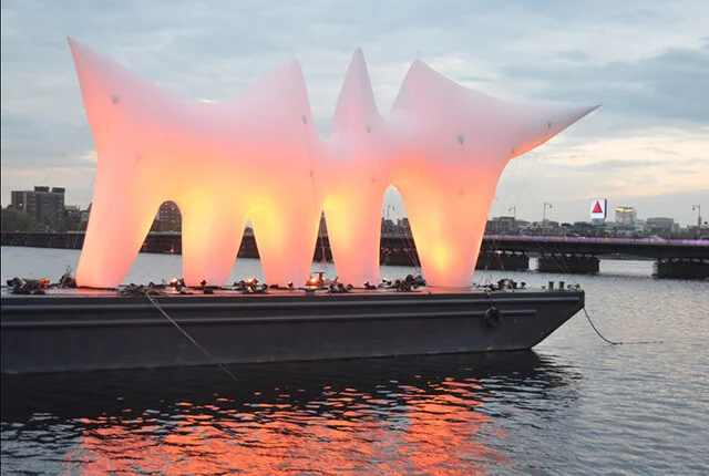 Large inflatable, abstract white sculpture glows orange and yellow as it sits on a floating platform in the water, with a cityscape and iconic Citgo sign visible in the background at dusk.