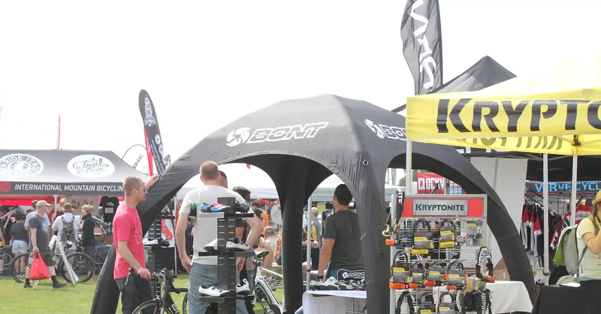 People are walking and browsing at an outdoor cycling expo with branded inflatable tents and booths, including Kryptonite and Bont, displaying bike locks and other products on a grassy area.