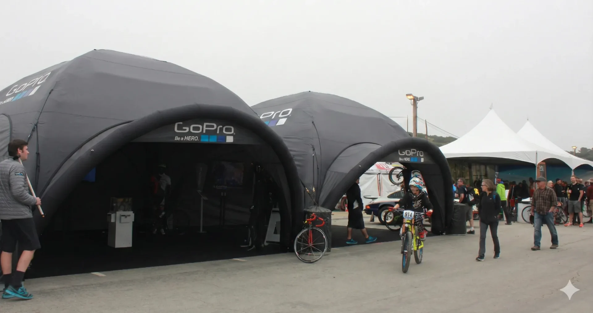 Two large black GoPro tents are set up outdoors with people walking and biking nearby. A person on a bicycle rides in front of the tents, while others gather and interact around the area. The sky is overcast.