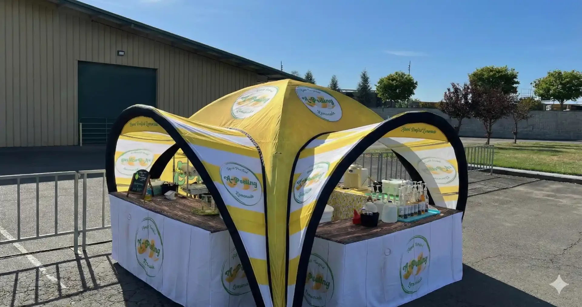 A yellow and white inflatable tent with branded logos covers a table displaying various beverages and syrups at an outdoor event. The setup is on pavement, with a metal fence and building in the background.