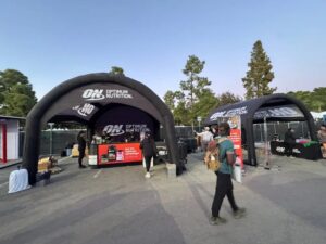 Two black Optimum Nutrition tents set up outdoors with tables displaying products. Several people are walking by or standing near the booths, and trees are visible in the background under a clear sky.