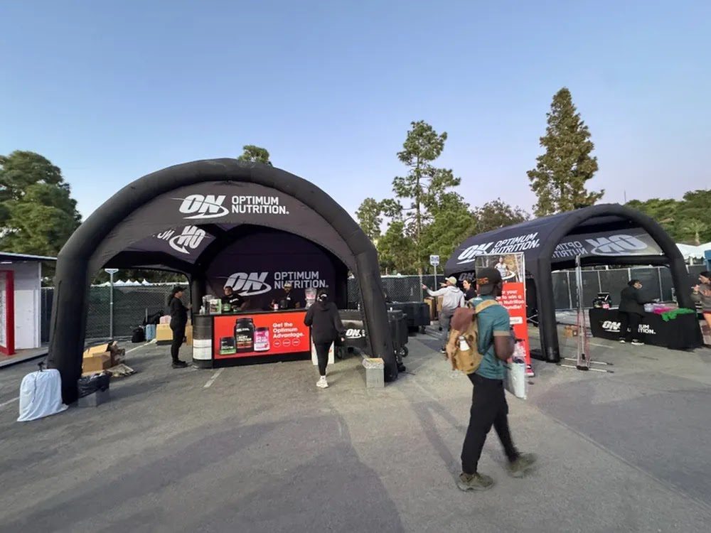 Two black Optimum Nutrition tents set up outdoors with tables displaying products. Several people are walking by or standing near the booths, and trees are visible in the background under a clear sky.