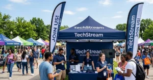 People gather at an outdoor tech fair, visiting booths under colorful tents. A group stands at the TechStream booth with banners reading JOIN US! on a sunny day, with trees in the background.